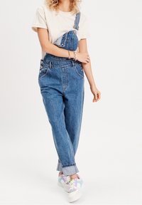 Pantalones de mezclilla con tirantes ajustables, bolsillo frontal y puños enrollados; combinados con zapatillas blancas con acentos en tonos pastel.