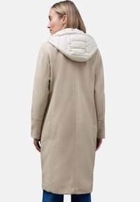 Beige Wollmischung knielanger Mantel mit einer gesteppte cremefarbenen Kapuze, ausgestattet mit seitlichen Taschen und einem geraden Schnitt. Glatte Textur und minimalistisches Design.