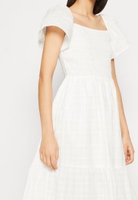 Robe blanche avec un décolleté carré, des manches bouffantes, un corsage texturé et une jupe plissée fluide avec un subtil motif en grille.