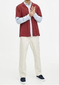 Chemise bordeaux à manches courtes portée sur une chemise à manches longues rayée bleu clair, associée à un pantalon crème et des chaussures à enfiler bleu foncé.