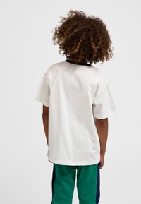 Göndör hajú gyermek hátat fordítva áll, laza fehér rövid ujjú inget és zöld nadrágot visel, melynek oldalán tengerészkék csíkok vannak, egyszínű háttér előtt.