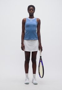 Γυναίκα που στέκεται με μπλε αμάνικο τοπ και λευκή φούστα, κρατώντας ρακέτα τένις στο πλευρό της σε απλό φόντο.