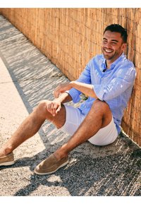 Lichtblauw overhemdshemd, witte shorts en beige instapschoenen. Man zit tegen een bamboeschutting, met zonlicht dat schaduwen creëert.