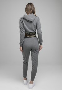 Grijze hoodie met ritssluiting en bijpassende joggers met een zwarte tailleband met gouden logo. Glad stof en geribbelde manchetten. Witte sneakers zichtbaar.