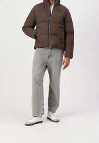 Hnědá péřová bunda na zip s kapucí, nošená přes šedé džíny se širokými nohavicemi a bílé tenisky se šedými detaily. Hladká, prošívaná textury.