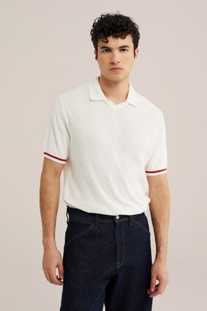 Giovane uomo con capelli ricci indossa una polo bianca a maniche corte in maglia con righe rosse sulle maniche e jeans blu scuro, in piedi davanti a uno sfondo semplice.
