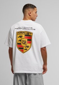 Hvid bomulds t-shirt med et stort gult og rødt skjoldgrafik på ryggen. Teksten "ULTIMATIV YDELSE" står over grafikken.