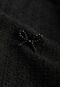Black fabric with a textured weave, featuring a black beaded embellishment shaped like a bow. The beads are faceted and shiny.
