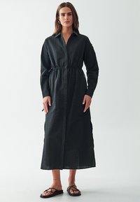 Abito nero a maniche lunghe con collo a camicia, vita cintrata, tessuto testurizzato, spacco laterale e chiusura con bottoni davanti.