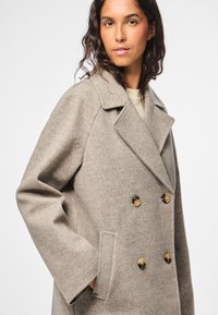 Cappotto di lana marrone chiaro doppiopetto con grandi bottoni dorati, rever a punta e tasche laterali, caratterizzato da un tessuto testurizzato.