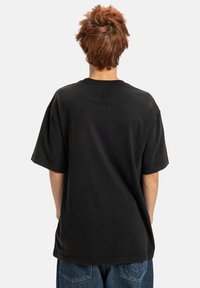 Person med kort, uordentlig rødligt hår iført en løs sort T-shirt og blå jeans, stående med ryggen til en ensfarvet baggrund.