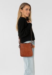 Borsa a tracolla in pelle intrecciata marrone con forma rettangolare, tracolla regolabile e pendente decorativo. Texture liscia e design strutturato.