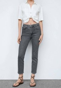 Jeans de mezclilla gris con cintura alta y ajuste ajustado, combinados con una camisa blanca corta de botones. Sandalias planas con tiras completan el conjunto.