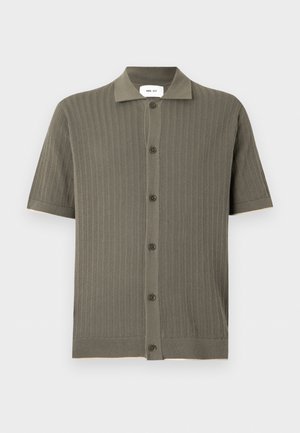 Mladý muž stojí s rukami za chrbtom, oblečený v olivovo zelenej krátkoruká blúzke na gombíky, béžových nohaviciach s širokými nohavicami a čiernych topánkach.