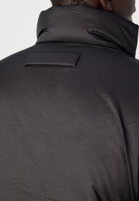 Homme portant une veste noire brillante à col montant, montrant l'arrière de l'épaule et la zone du cou avec un détail de patch rectangulaire.