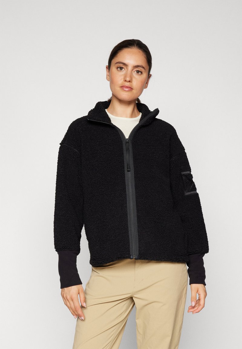 Giacca in pile nera con zip completa, colletto alto e vestibilità oversize. Presenta maniche lunghe con polsini neri e una toppa con logo laterale.