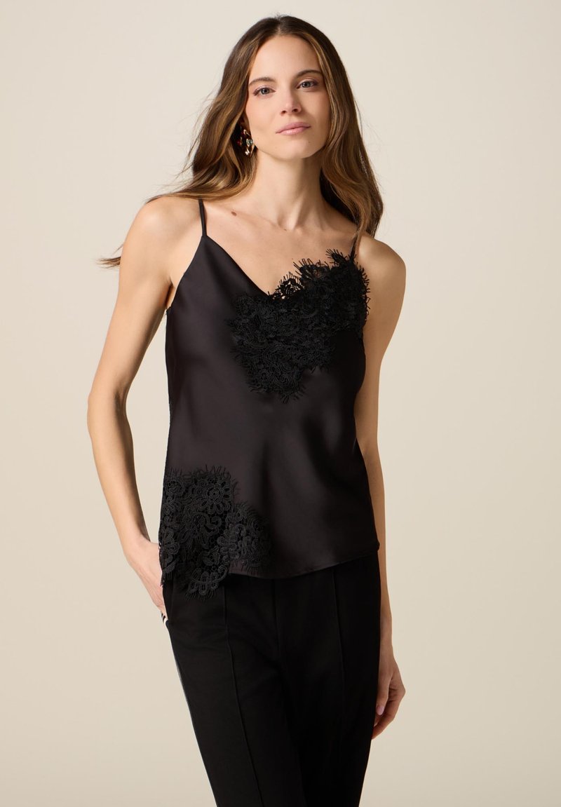 Kvinde iført sort satin cami med blonder og sorte bukser, stående mod beige baggrund med den ene hånd i lommen.