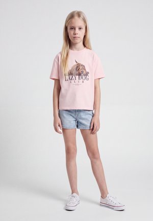 Lyshåret pige står iført en pink T-shirt med hundegrafik og tekst "Lazy Dog Club,", lyseblå denimshorts og hvide sneakers på en ensfarvet baggrund.