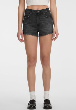 Nő magas derekú fekete farmer rövidnadrágban, kopott szélű, fehér zokniban, fekete mokaszinban és egy rövidített fehér ujjatlan felsőben.