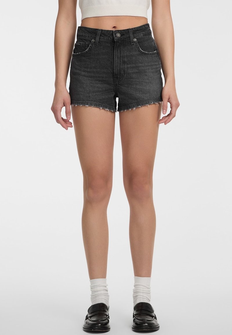 Naine, kes kannab kõrge vöökohaga musti teksapükse franjeeritud äärtega, valgeid sokke, musti mokasiine ja kärbitud valget varrukateta topi.