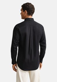 Homme de dos portant une chemise noire à manches longues avec des poignets boutonnés et un pantalon blanc sur un fond uni.