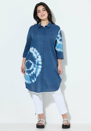 Kvinde iført en knælængde blå slå-om-kjole med hvide batikcirkler, hvide bukser og sølvfarvede sandaler med spænde, stående foran en hvid baggrund.