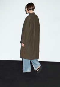 Camicia giacca oversized verde oliva con maniche lunghe, fessure laterali e colletto classico, abbinata a jeans a gamba larga blu chiaro e sandali.