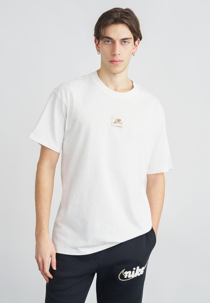 Fehér pamut rövid ujjú póló, amelyen egy kis hímzett logó látható a mellrészen. Fekete melegítőnadrággal párosítva, amelyen szintén logó található.