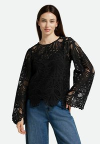 Top in pizzo nero con maniche lunghe, caratterizzato da motivi intricati e orlo scallop. Abbinato a pantaloni in denim blu.