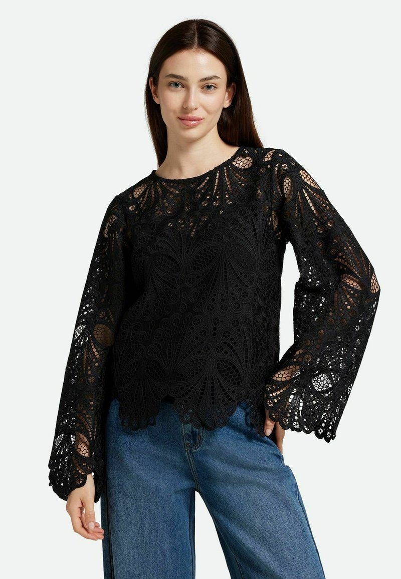 Top in pizzo nero con maniche lunghe, caratterizzato da motivi intricati e orlo scallop. Abbinato a pantaloni in denim blu.