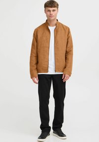 Veste en daim brun avec un col montant, fermeture éclair et poches latérales, associée à une chemise blanche et un pantalon noir, portée par une personne.