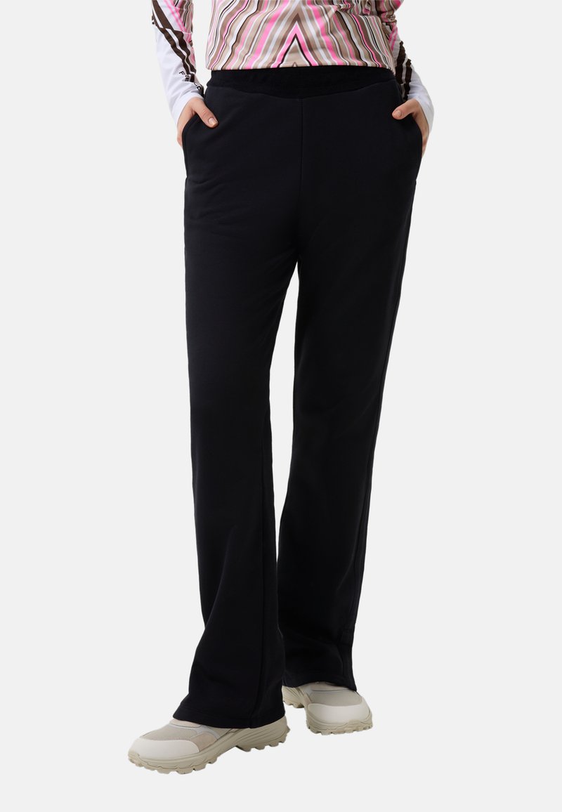 Black, relaxed-fit pants with an elastic waistband, featuring side pockets and a smooth texture, paired with light-colored athletic shoes.