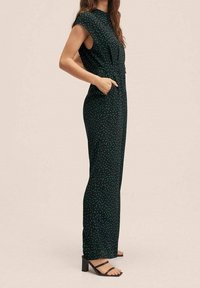 Zelený overal s puntíky, vysokým výstřihem, bez rukávů, bočními kapsami a širokými nohavicemi, doplněný černými sandály na vysokém podpatku.