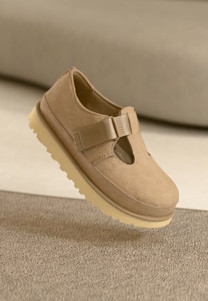 Tan suede shoe with a rounded toe, adjustable strap, and rubber sole featuring a textured pattern, hovering above a surface.