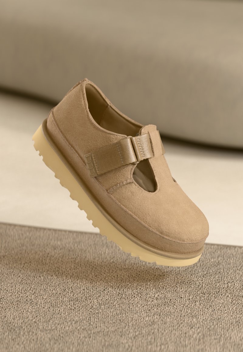 Tan suede schoen met een ronde neus, verstelbare band en rubberen zool met een getextureerd patroon, zwevend boven een oppervlak.