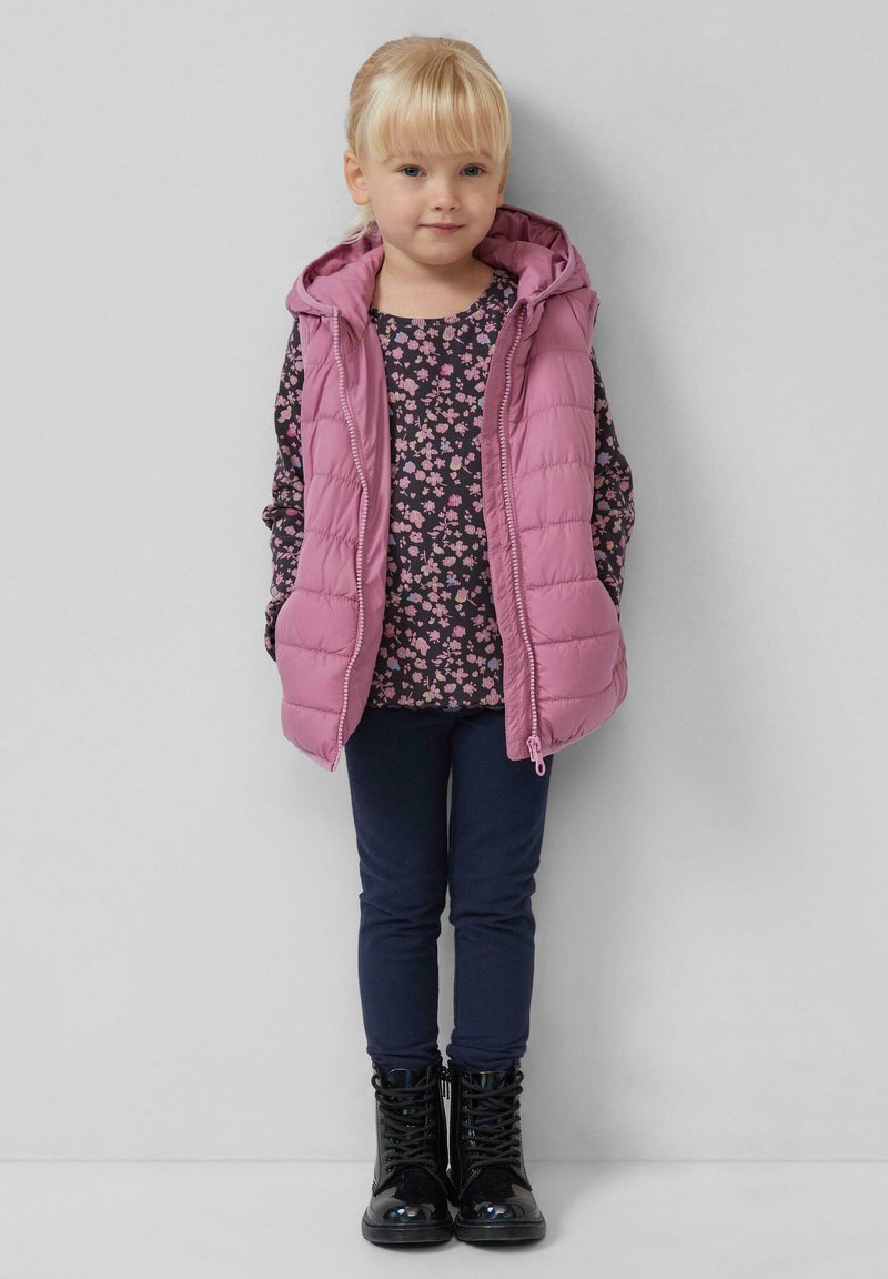 Roze gewatteerde vest met capuchon over een donkerbloemig shirt, gecombineerd met marineblauwe legging en glanzende zwarte laarzen. Soepele textuur en praktische vormgeving.