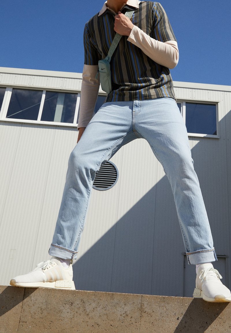 Personne portant un jean bleu clair, des baskets blanches, une chemise à manches courtes rayée par-dessus une chemise beige à manches longues, debout, jambes écartées, à l'extérieur près d'un bâtiment.