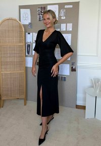 Fekete, testhez simuló ruha mély V-kivágással, fodros ujjal és combig nyílással. Fekete, hegyes orrú magassarkú cipővel viselve, tervezői vázlatok előtt.