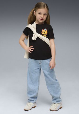 Jong meisje met lang blond haar draagt een zwart T-shirt met een kattenafbeelding, lichtblauwe spijkerbroek, witte sneakers en een crèmekleurige sweatshirt over haar schouders.