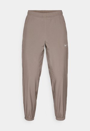 Nike athletic pants in a light brown color, featuring an elastic waistband, tapered legs, and side pockets. Smooth, lightweight fabric.