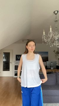 Biały, luźny top na ramiączkach nałożony na dopasowaną białą koszulę. Niebieskie spodnie sportowe z białymi paskami, ustawione w jasnym pomieszczeniu.