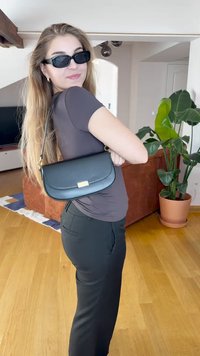 Borsa a tracolla in pelle nera con design piatto, chiusura dorata e tracolla regolabile. Il modello indossa un top aderente e pantaloni scuri, in un ambiente interno.