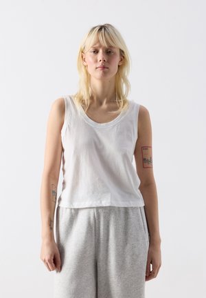 Blonde vrouw met een witte mouwloze top en grijze joggingbroek, staand tegen een effen witte achtergrond met zichtbare armtattoeages.