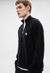 Čierny pletený pulóver s vysokým golierom a polovičným zipsom. Obsahuje malú vyšívanú nášivku s logom. Mäkká textúra s voľným strihom.
