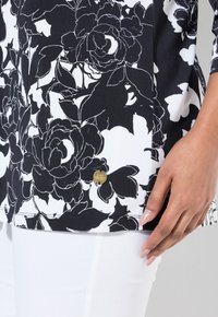 Schwarzes und weißes Blumenshirt mit glatter Textur, das große Blütenmuster zeigt. Ein kleines goldenes Akzentetikett ist in der Nähe des Saums sichtbar.