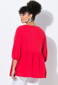 Top rosso con texture, maniche a tre quarti, orlo svasato e scollatura rotonda, mostrato dal retro su uno sfondo bianco.