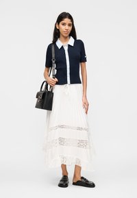 Γυναίκα που φοράει μπλε ναυτικό κοντομάνικο πουκάμισο με κουμπιά, λευκή δαντελένια φούστα, μαύρα μοκασίνια και κρατάει μια μαύρη τσάντα στον ώμο της.