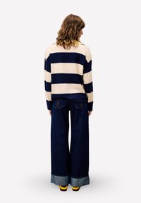 Persoon met golvend bruin haar, gekleed in een crèmekleurige en marineblauwe gestreepte trui, wijdvallende donkere jeans met omslagen, en gele schoenen, met de rug naar de kijker.