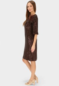 Brązowa sukienka z cekinami, z rękawami do połowy długości i piórowym akcentem na ramieniu, noszona z beżowymi butami na obcasie na jednolitym tle.