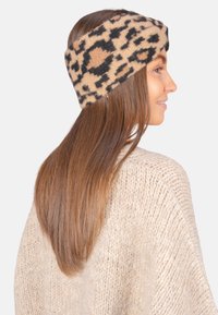 Bandeau en tissu imprimé léopard beige et noir, texture douce, design large couvrant le front, porté avec de longs cheveux bruns et un pull léger.